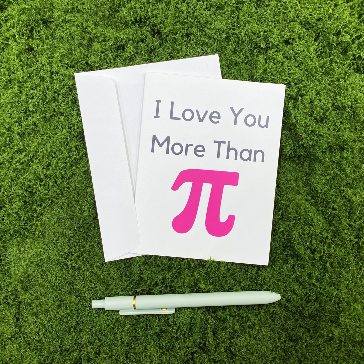 Folded greeting card reading "I Love You More Than" with a pink pi symbol, resting on a white envelope on a mossy surface, with a sage green pen below.