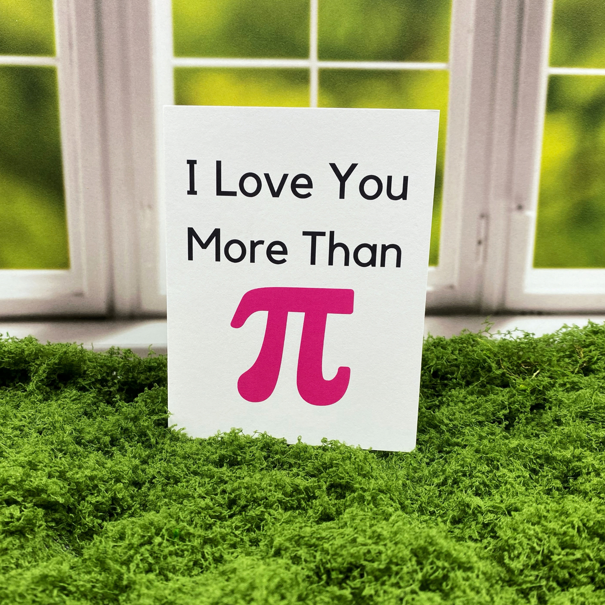 Greeting card that reads "I Love You More Than" in black text above a large pink pi symbol, standing upright on a moss surface in front of a white-framed window.