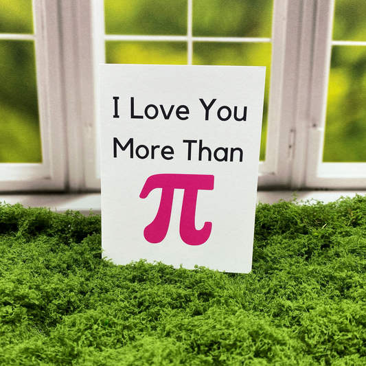 Greeting card that reads "I Love You More Than" in black text above a large pink pi symbol, standing upright on a moss surface in front of a white-framed window.