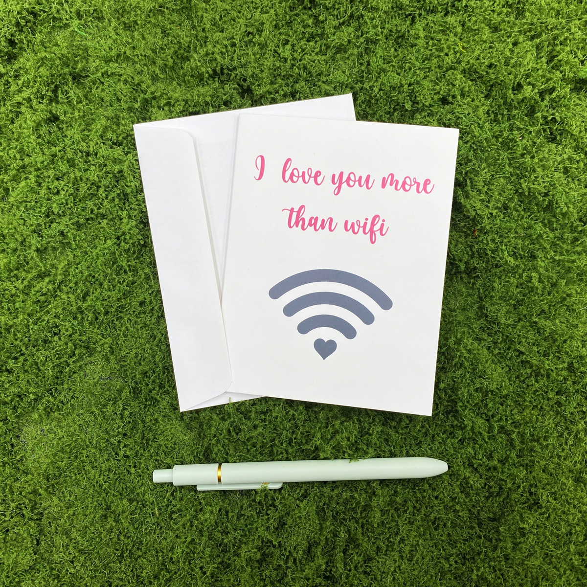 Folded greeting card reading "I love you more than wifi" lying on a white envelope on a mossy surface, with a sage green pen resting below.