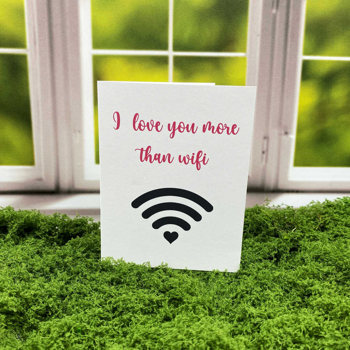Greeting card that reads "I love you more than wifi" in pink script above a black wifi symbol with a heart as the bottom signal bar, standing upright on a moss surface in front of a white-framed window.