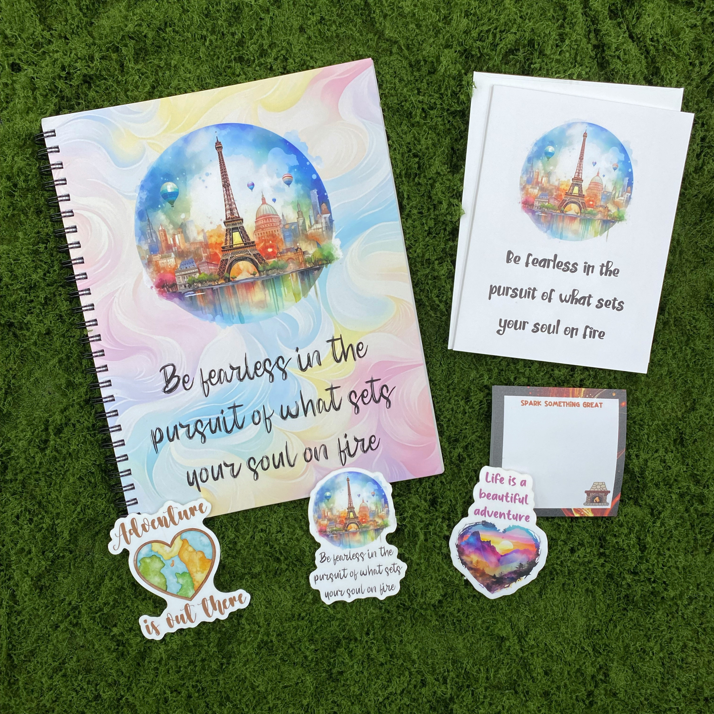 A motivational gift set including a be fearless spiral notebook, a matching greeting card with envelope, a spark something great sticky note pad, and three watercolor die-cut stickers featuring a world map heart, Eiffel Tower scene, and mountain sunset heart, arranged on a green moss surface