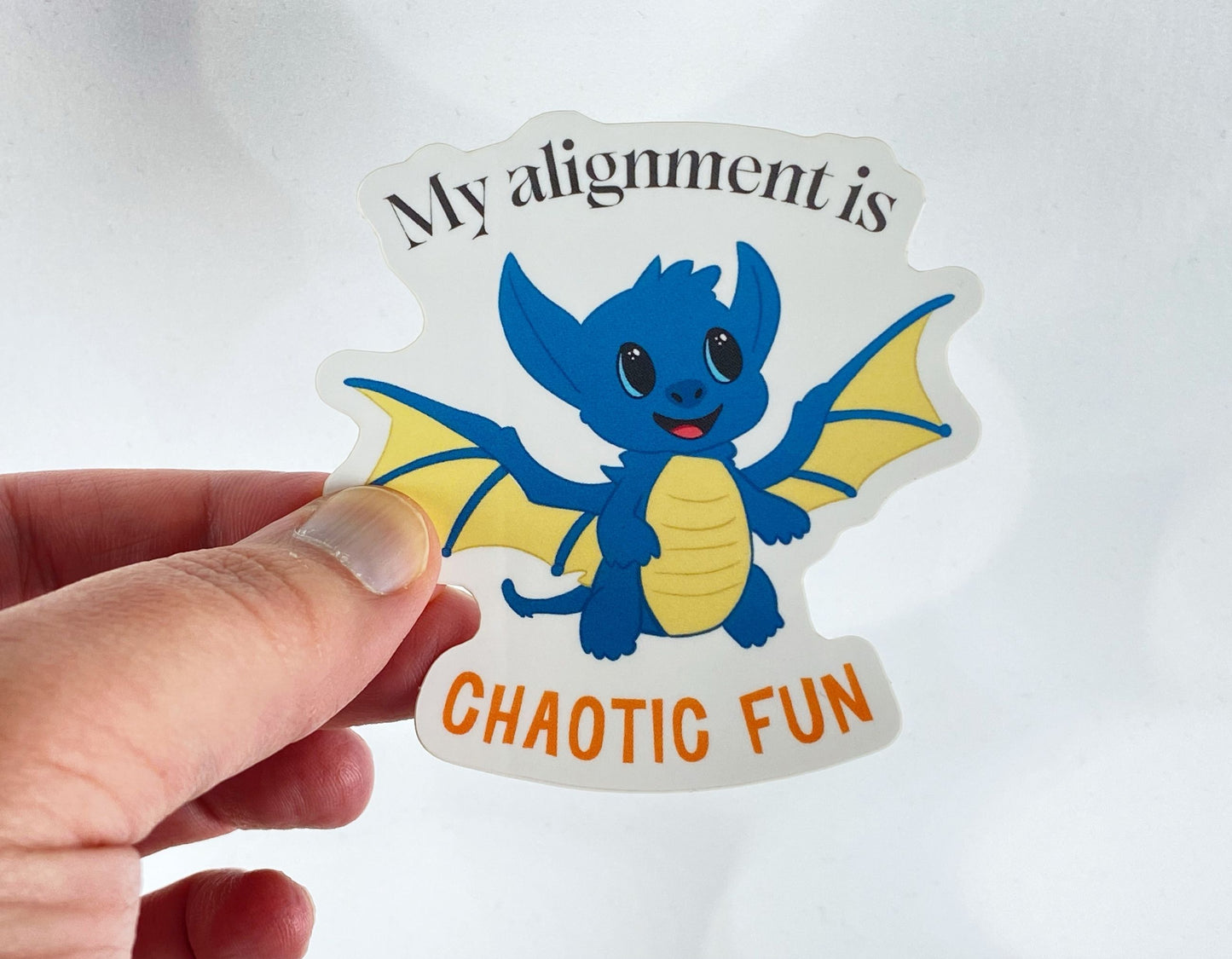 Die-cut vinyl sticker held between fingers, showing a smiling blue baby dragon with yellow wings, above the text "My alignment is chaotic fun," on a white background