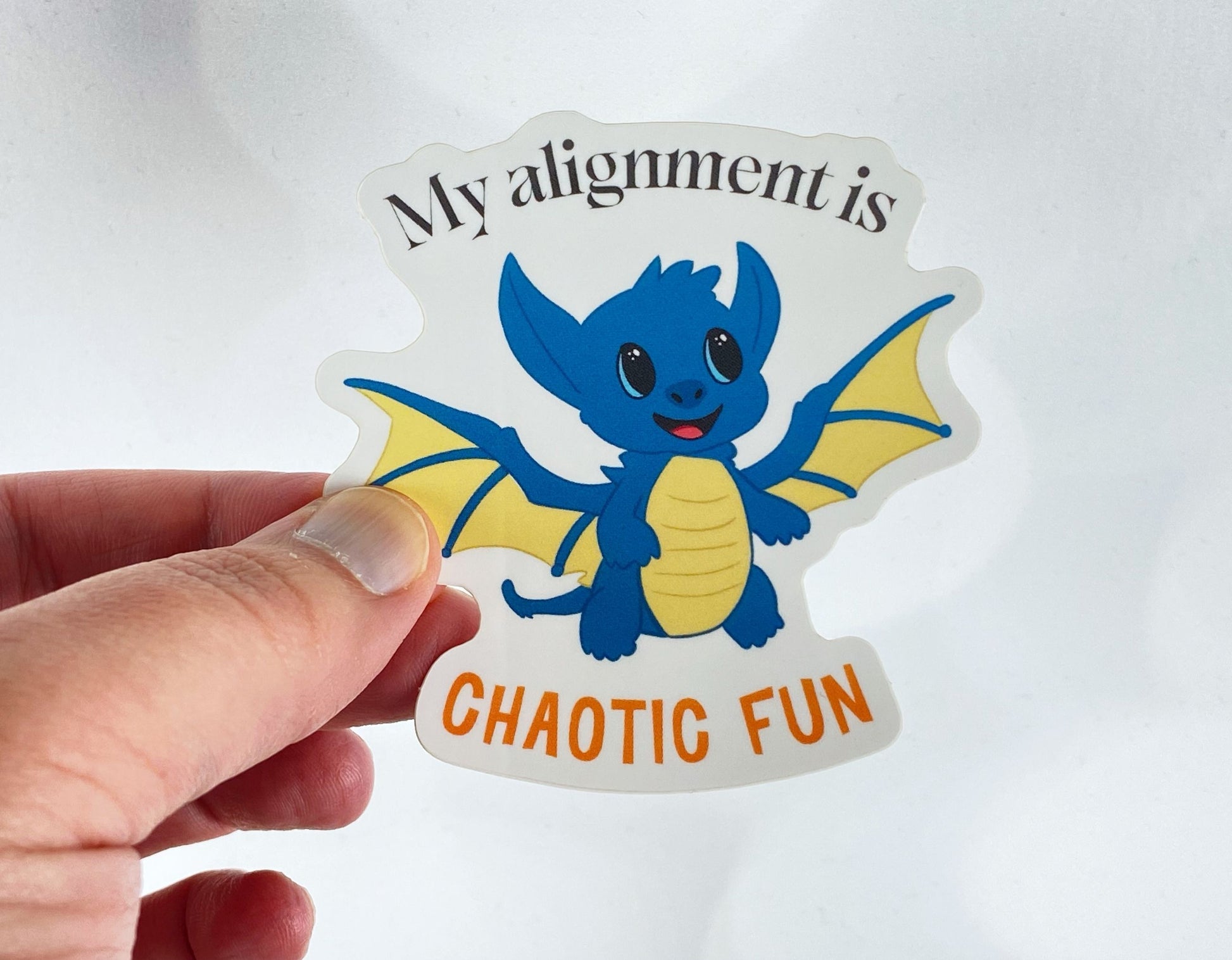 Die-cut vinyl sticker held between fingers, showing a smiling blue baby dragon with yellow wings, above the text "My alignment is chaotic fun," on a white background
