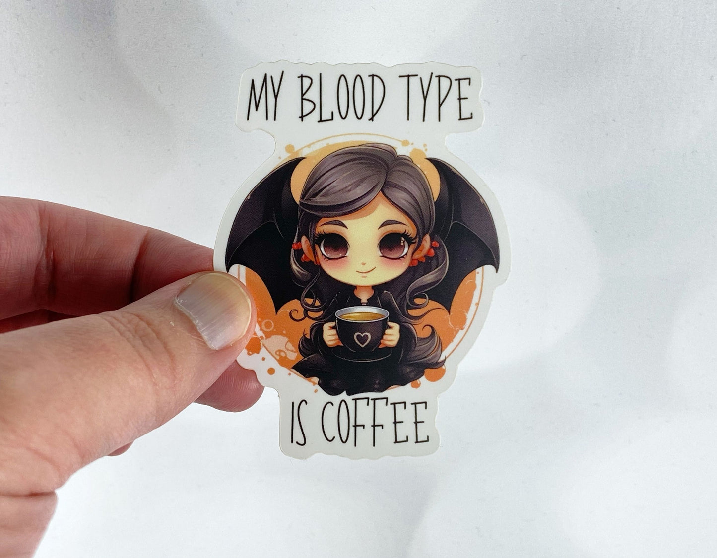 A hand holds a sticker illustration of a bat-winged girl holding a mug of coffee, reading "My blood type is coffee," against a white background.