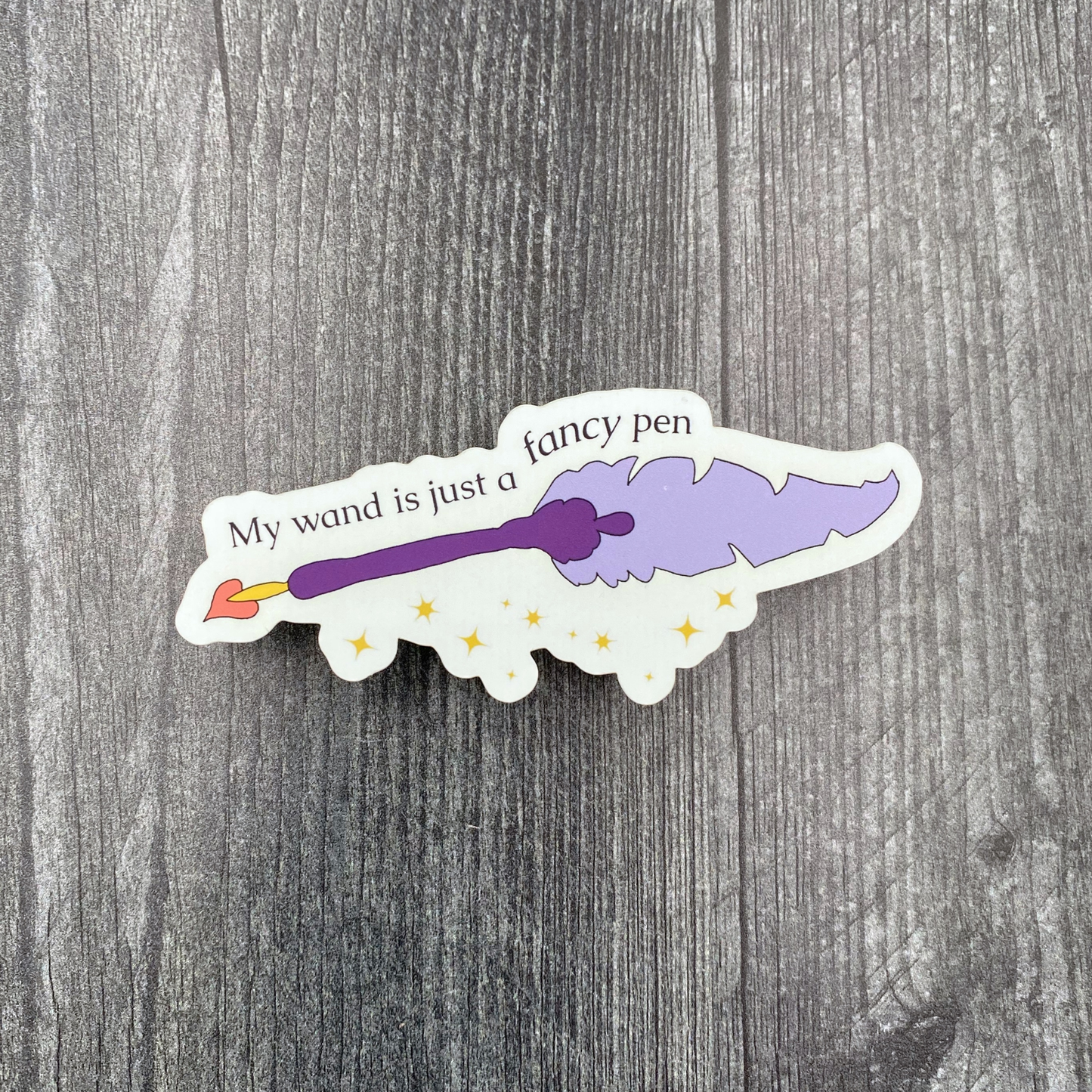 Die-cut vinyl sticker showing a sparkling purple quill pen with a hand-drawn heart, with the text "My wand is just a fancy pen," on a gray wood background