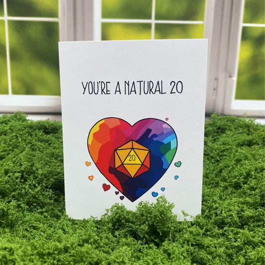 "You're a Natural 20" greeting card standing upright on a moss mat in front of a window backdrop, featuring a rainbow heart with a 20-sided die and small hearts around it.
