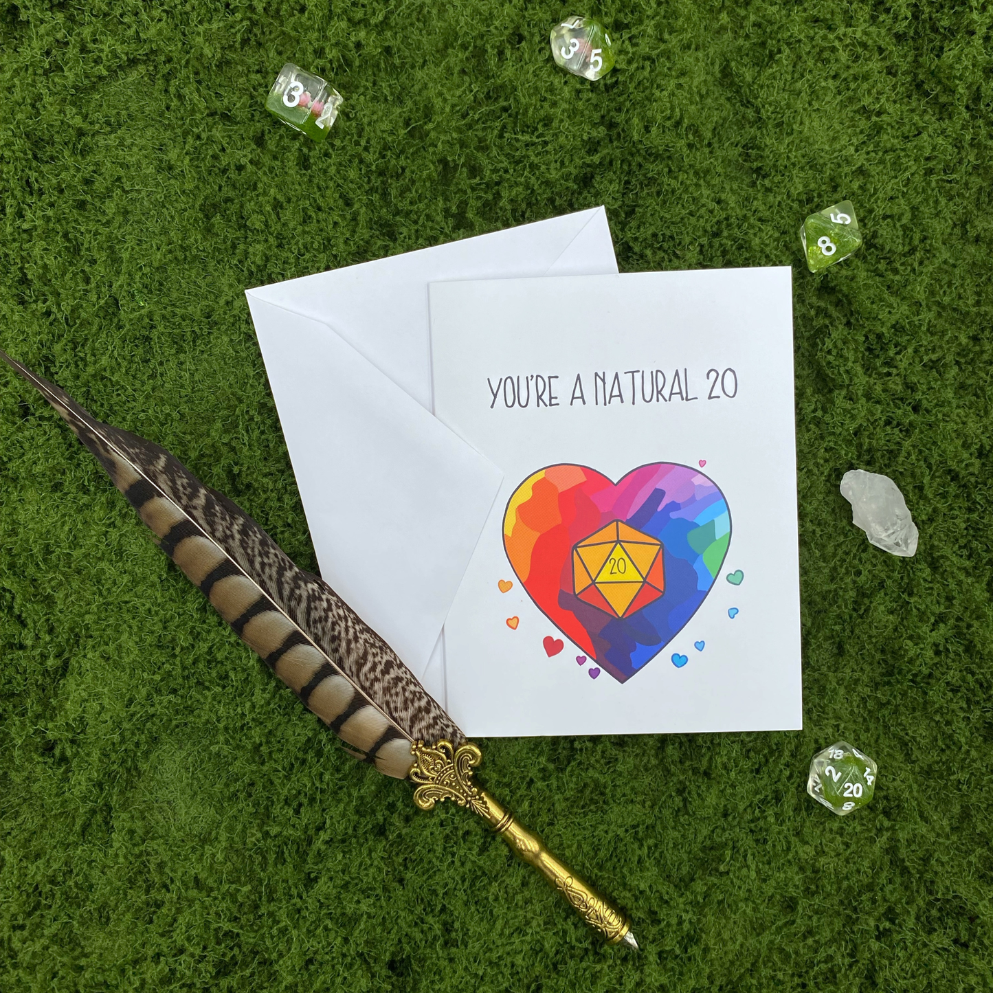 Greeting card that reads "You're a Natural 20" with a rainbow heart and gold twenty-sided die design, propped on its envelope, surrounded by dice and a feather quill pen on a grass background.