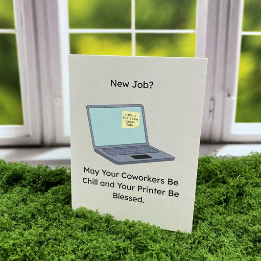 Greeting card that reads "New Job?" above an illustration of a laptop with a sticky note reading "CTRL + ALT + New Career Path!" on the screen, and the text "May Your Coworkers Be Chill and Your Printer Be Blessed" below, standing upright on a moss surface in front of a white-framed window.