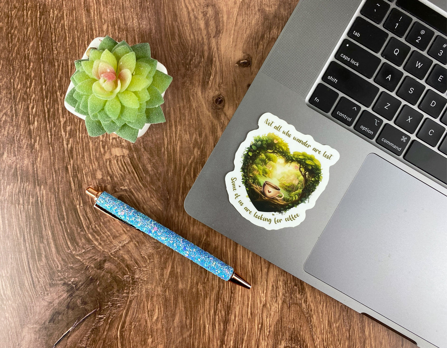 Heart-shaped forest and coffee cup sticker applied to the corner of a silver laptop trackpad, next to a succulent and a glitter pen on a wood desk
