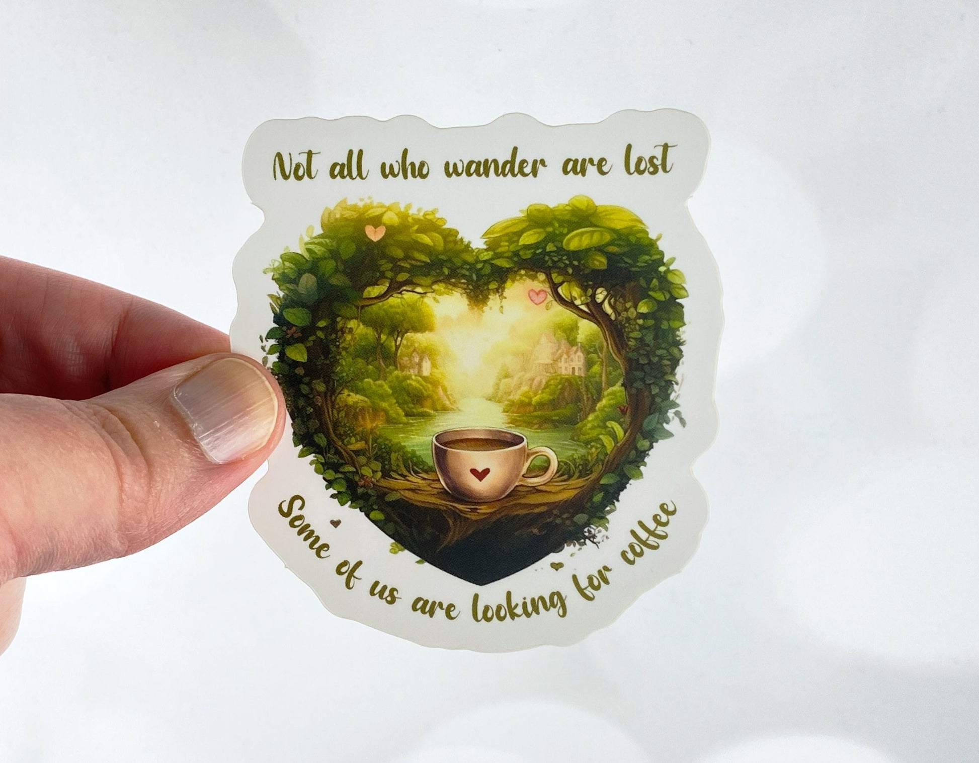 Die-cut vinyl sticker held between fingers, showing a heart-shaped forest scene with a coffee cup at its center, with the text "Not all who wander are lost, some of us are looking for coffee," on a white background