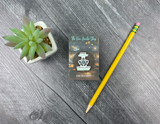 Enamel pin shaped like a shield crest with a d20 in the center, reading "I'm not clumsy, I rolled low on dexterity," on a backing card next to a succulent and pencil on a gray wood background
