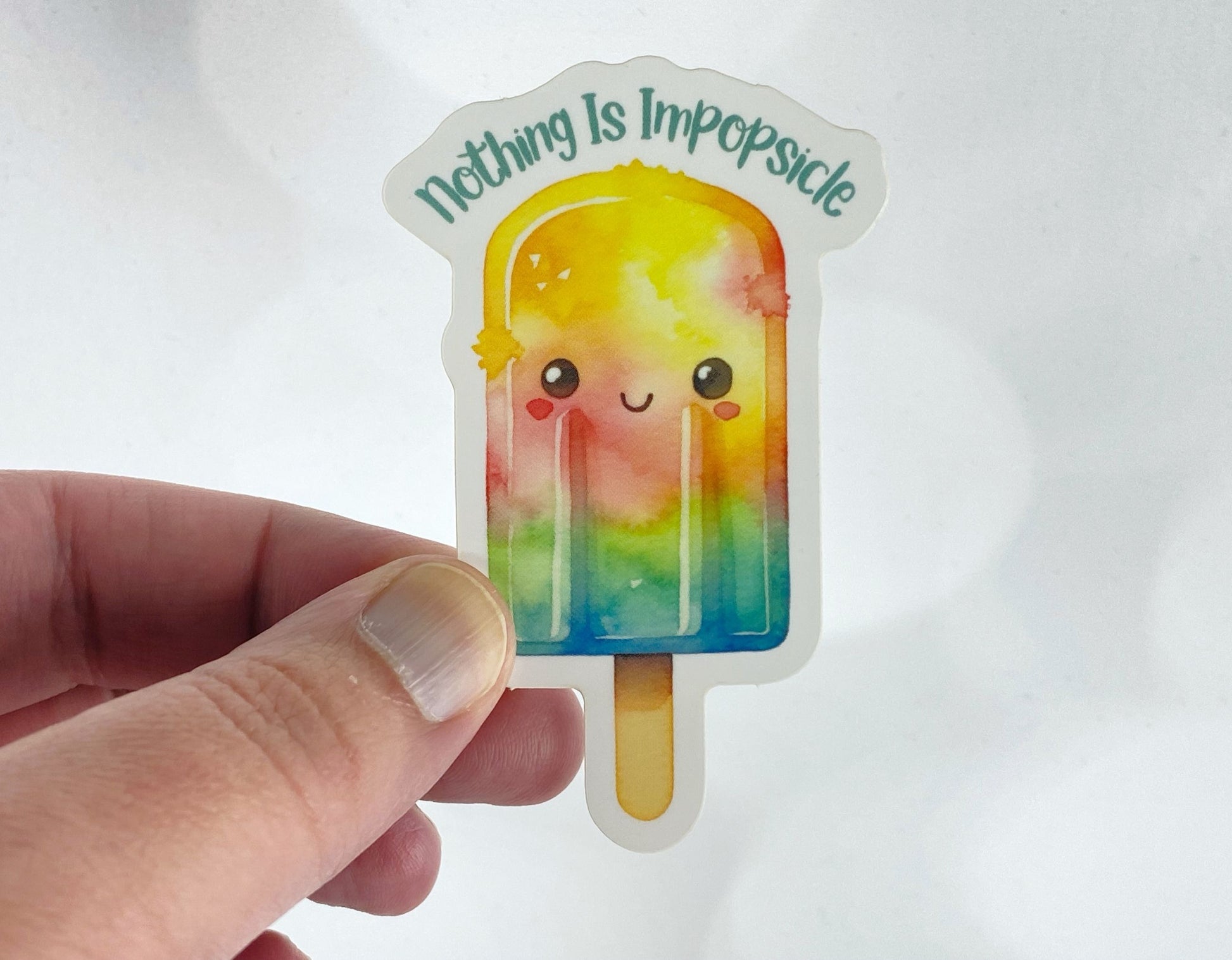 A hand holds a sticker illustration of a smiling watercolor popsicle, reading "Nothing is impopsicle," against a white background.