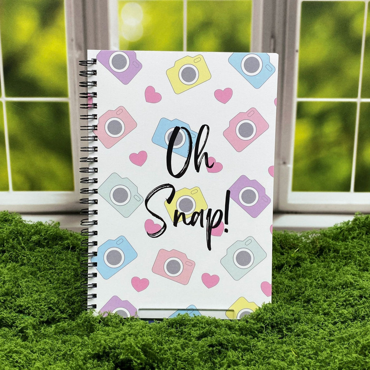 Spiral-bound notebook with a pastel pattern of cameras and hearts, reading 'Oh Snap!' in black script, displayed on a moss mat with a window backdrop.