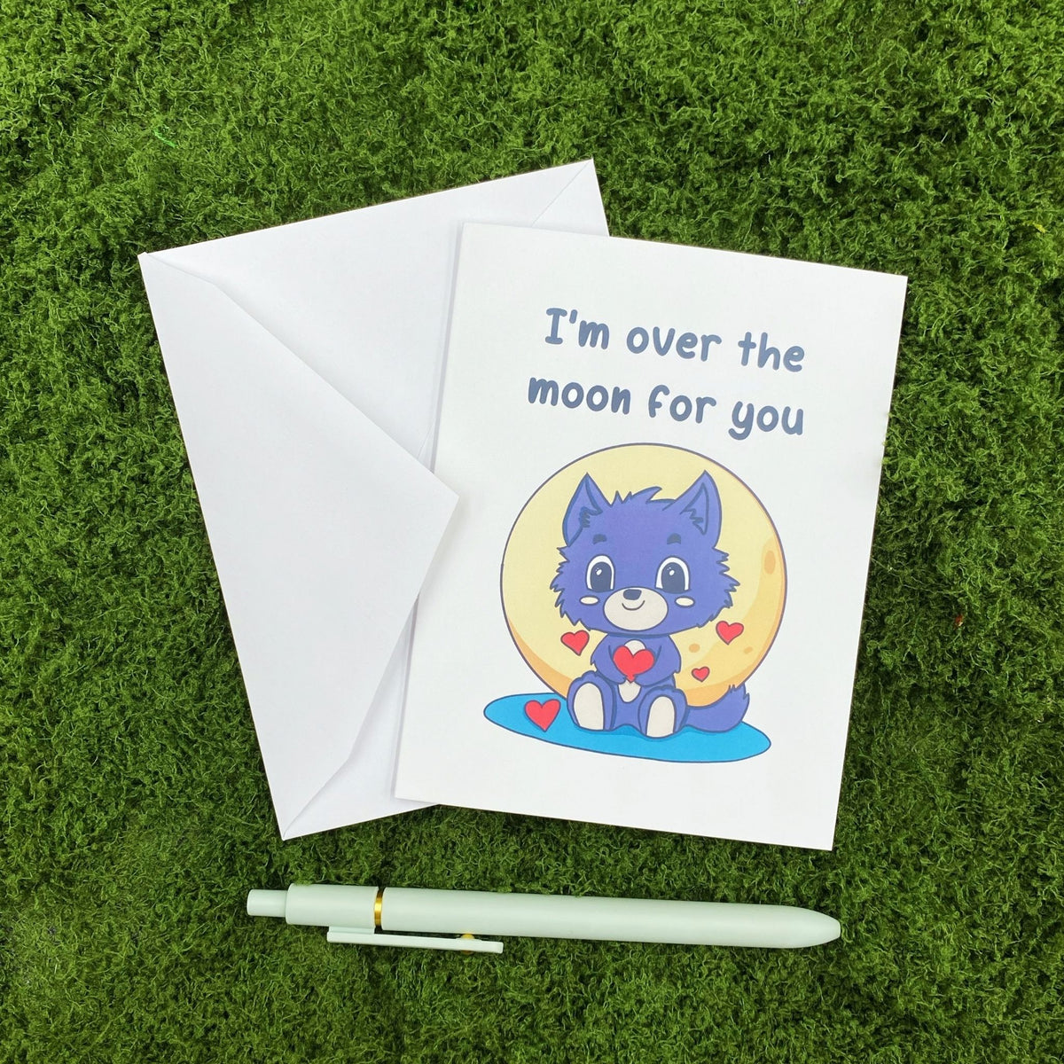 "I'm Over the Moon for You" greeting card lying on a white envelope on grass, with a mint green pen beside it for scale.