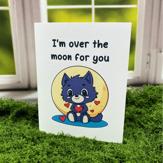 "I'm Over the Moon for You" greeting card standing upright on a moss mat in front of a window backdrop, featuring an illustrated baby werewolf holding a heart in front of a full moon, surrounded by small hearts.