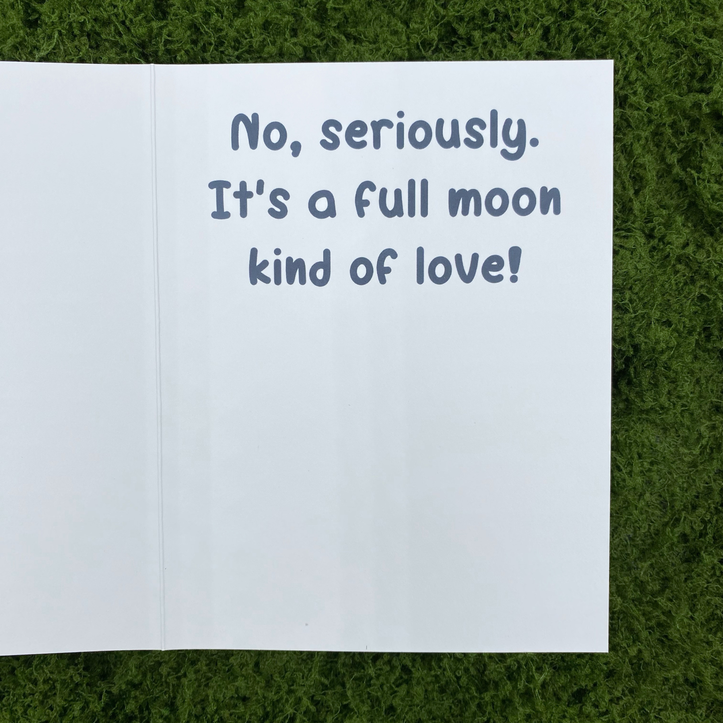 Inside spread of the greeting card showing the printed message "No, seriously. It's a full moon kind of love!" lying open on artificial grass.