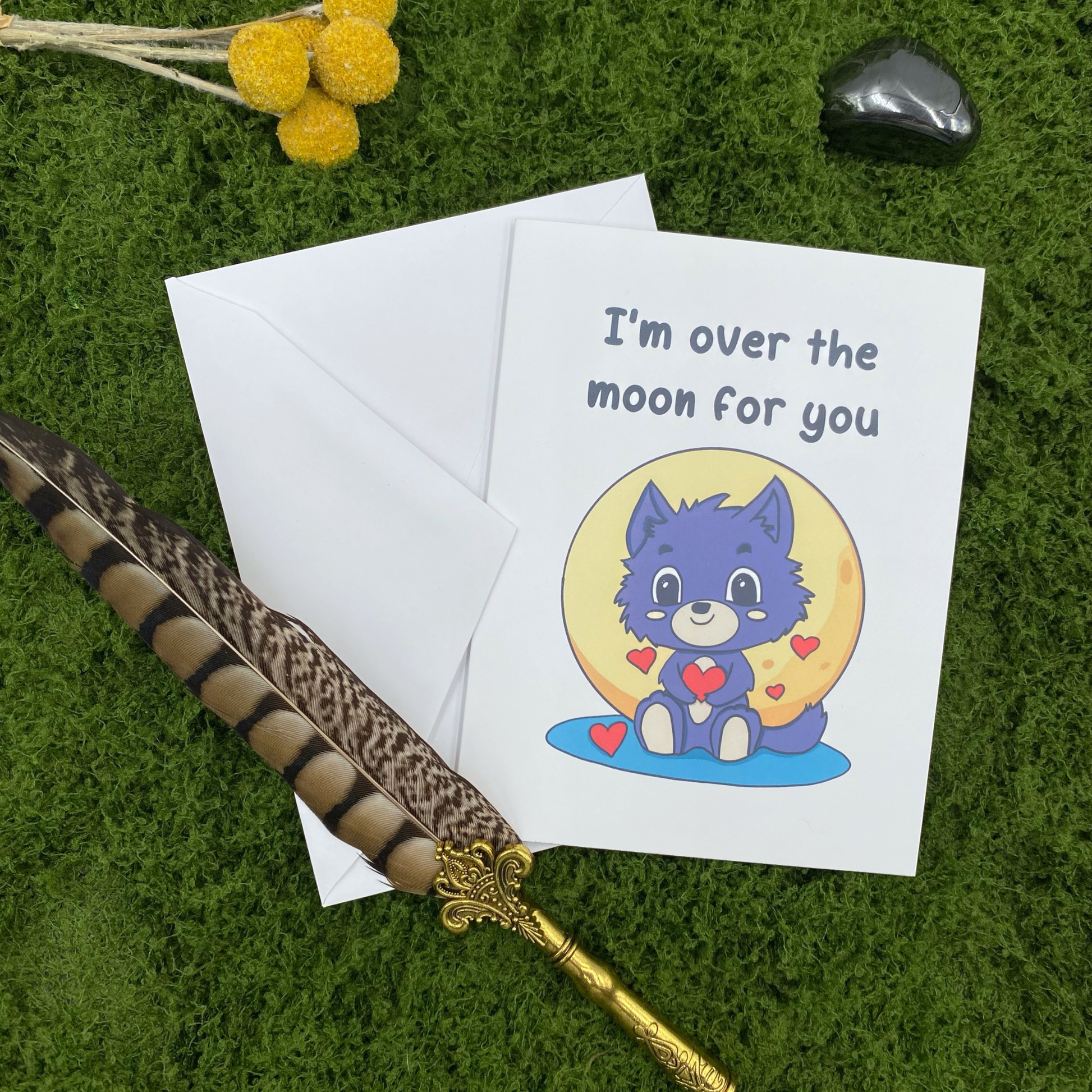 Greeting card that reads "I'm over the moon for you" with an illustration of a grey werewolf pup holding a heart in front of a glowing moon, propped against its envelope on artificial grass with a feather quill pen, dried flowers, and an obsidian stone.