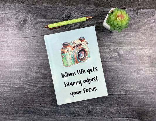 Hardcover journal in pale blue with an illustrated vintage camera and the words "When life gets blurry, adjust your focus," resting on a grey wood surface next to a glitter pen and small succulent.