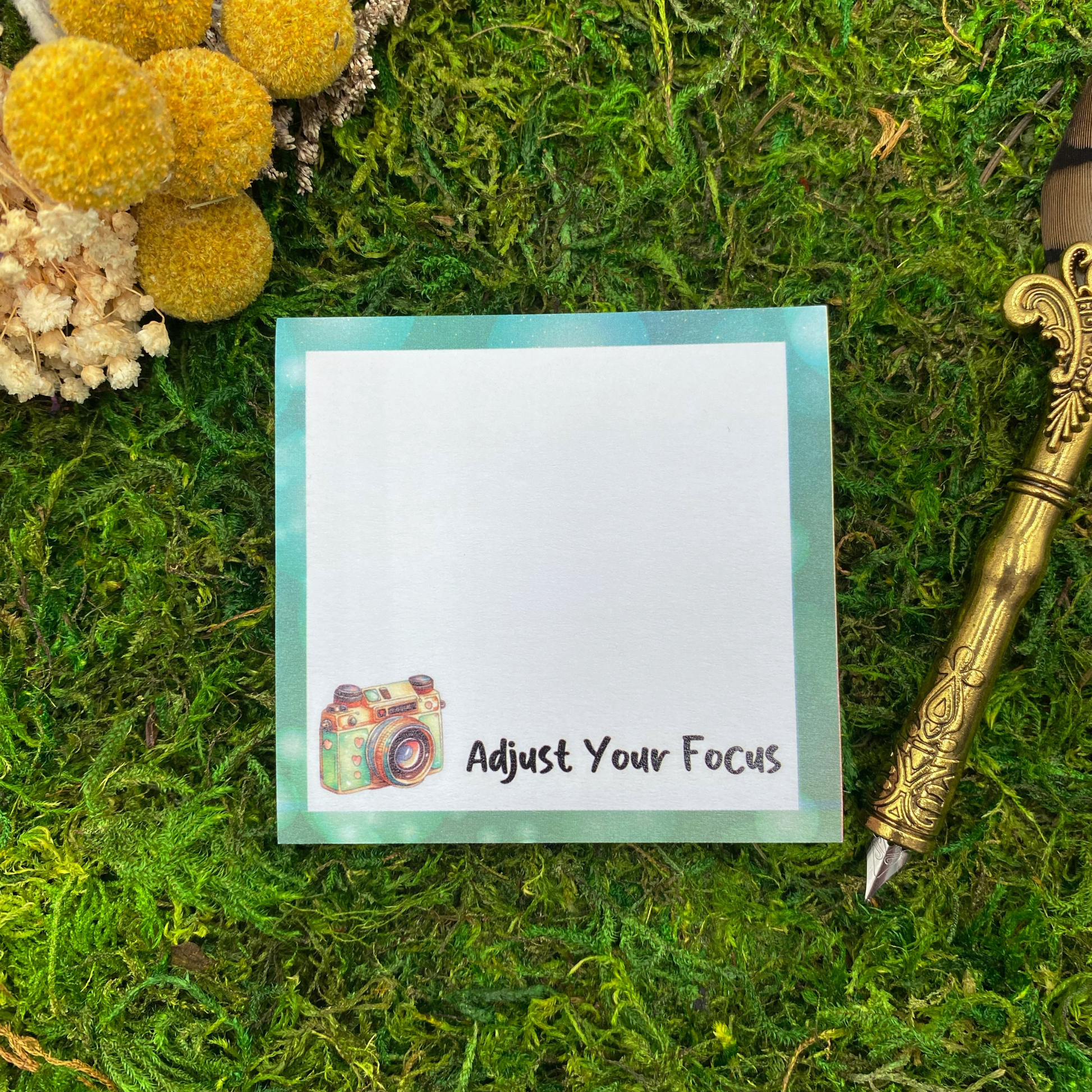 Square sticky note pad with a teal border, an illustrated vintage camera, and the words "Adjust Your Focus," resting on moss next to dried yellow and white flowers and a gold quill pen.