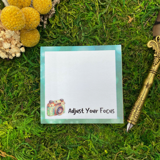 Square sticky note pad with a teal border, an illustrated vintage camera, and the words "Adjust Your Focus," resting on moss next to dried yellow and white flowers and a gold quill pen.