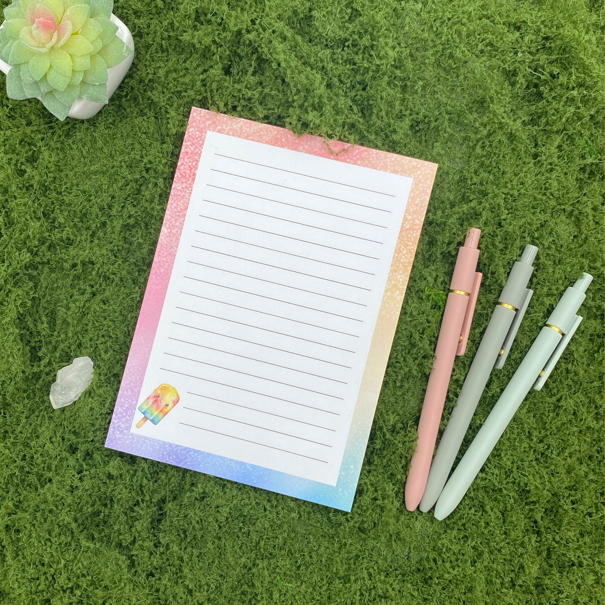Lined notepad with a pastel rainbow watercolor border and a smiling popsicle illustration in the corner, resting on artificial grass next to a crystal, small succulent, and three pastel gel pens.