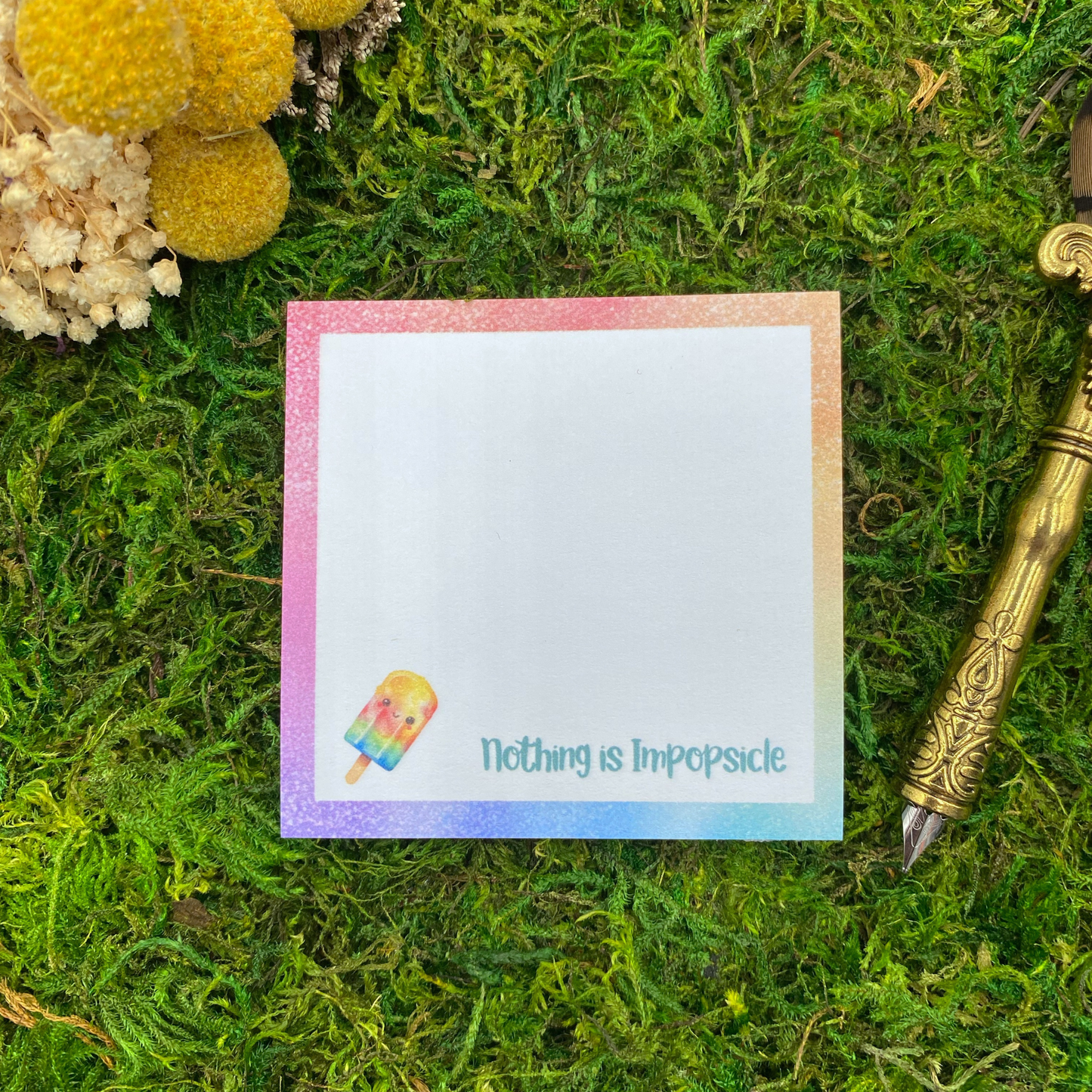 Square sticky note pad with a pastel rainbow border, a smiling popsicle illustration, and the words "Nothing Is Impopsicle," resting on moss next to dried yellow and white flowers and a gold quill pen.