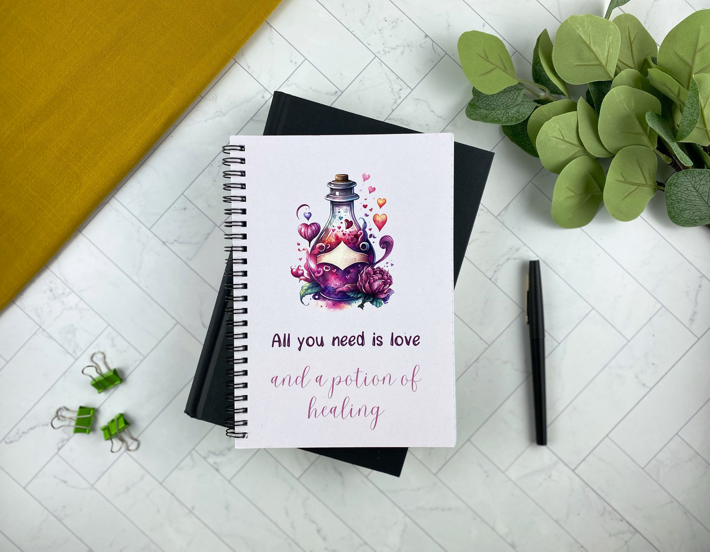Spiral notebook cover illustrated with a glowing potion bottle surrounded by hearts and pink flowers, reading "All you need is love and a potion of healing," resting on a black notebook with green binder clips, a black pen, and eucalyptus greenery nearby.