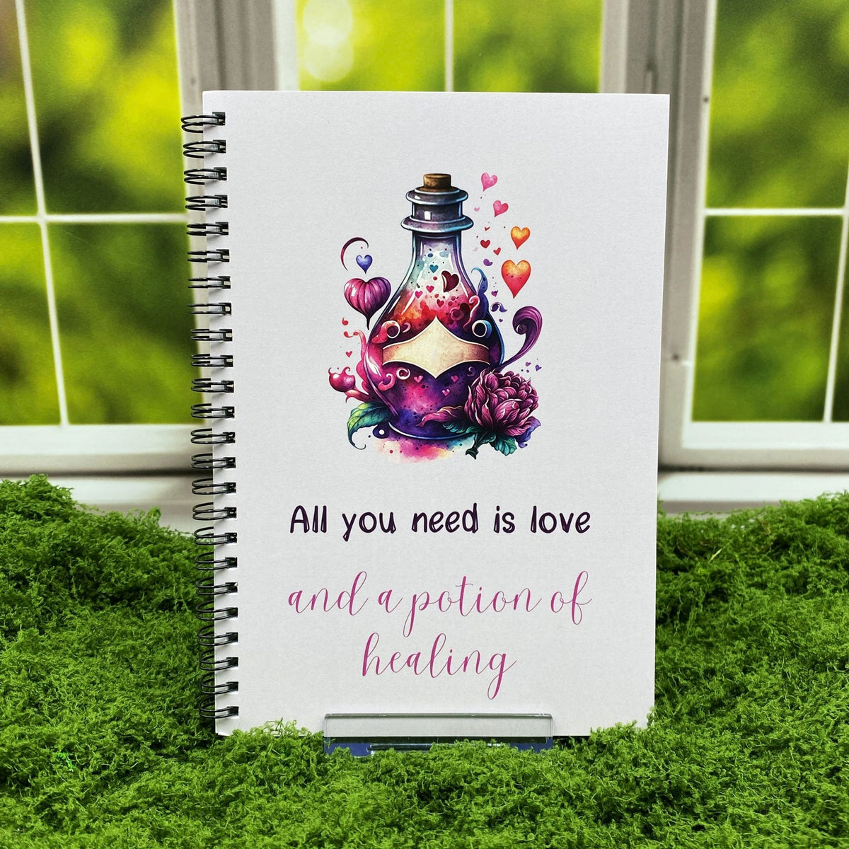 Spiral-bound notebook with a watercolor potion bottle illustration surrounded by hearts and flowers, reading 'All you need is love and a potion of healing,' displayed on a moss mat with a window backdrop.