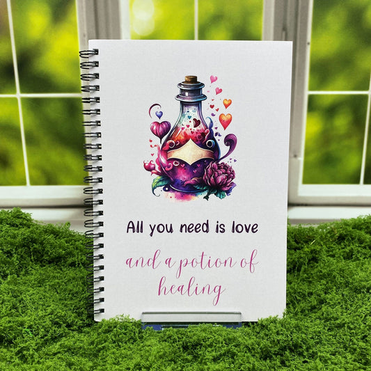 Spiral-bound notebook with a watercolor potion bottle illustration surrounded by hearts and flowers, reading 'All you need is love and a potion of healing,' displayed on a moss mat with a window backdrop.