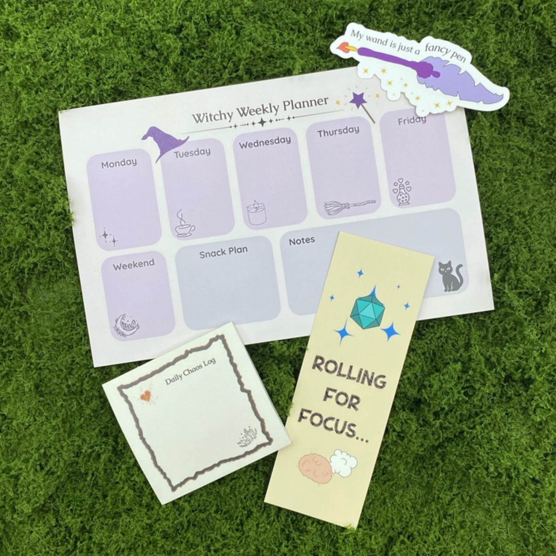 A curated gift set on artificial grass featuring a "Witchy Weekly Planner" notepad with witch hat and cauldron illustrations, a "Daily Chaos Log" sticky note, a "Rolling for Focus" bookmark with a teal d20, and a quill pen sticker reading "My wand is just a fancy pen."
