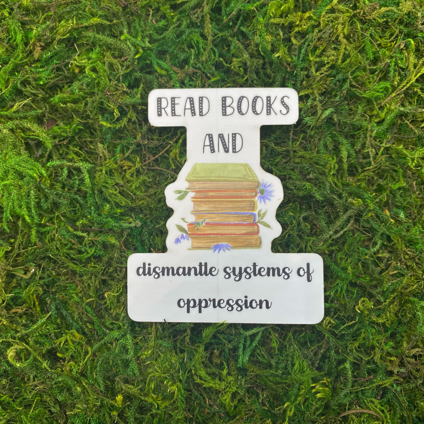 A sticker reading "Read books and dismantle systems of oppression" with an illustration of a stack of books surrounded by blue wildflowers, resting on artificial moss.