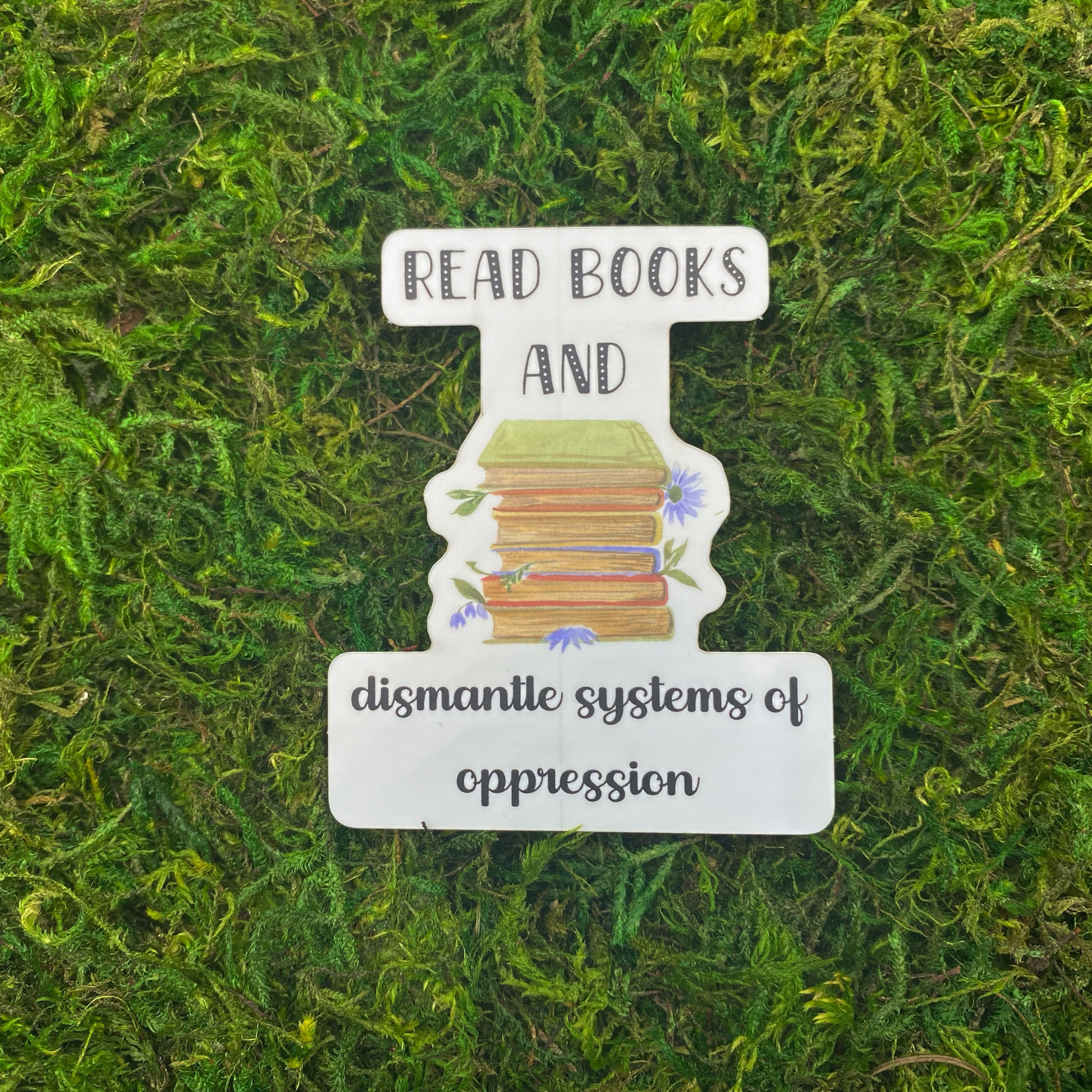 A sticker reading "Read books and dismantle systems of oppression" with an illustration of a stack of books surrounded by blue wildflowers, resting on artificial moss.