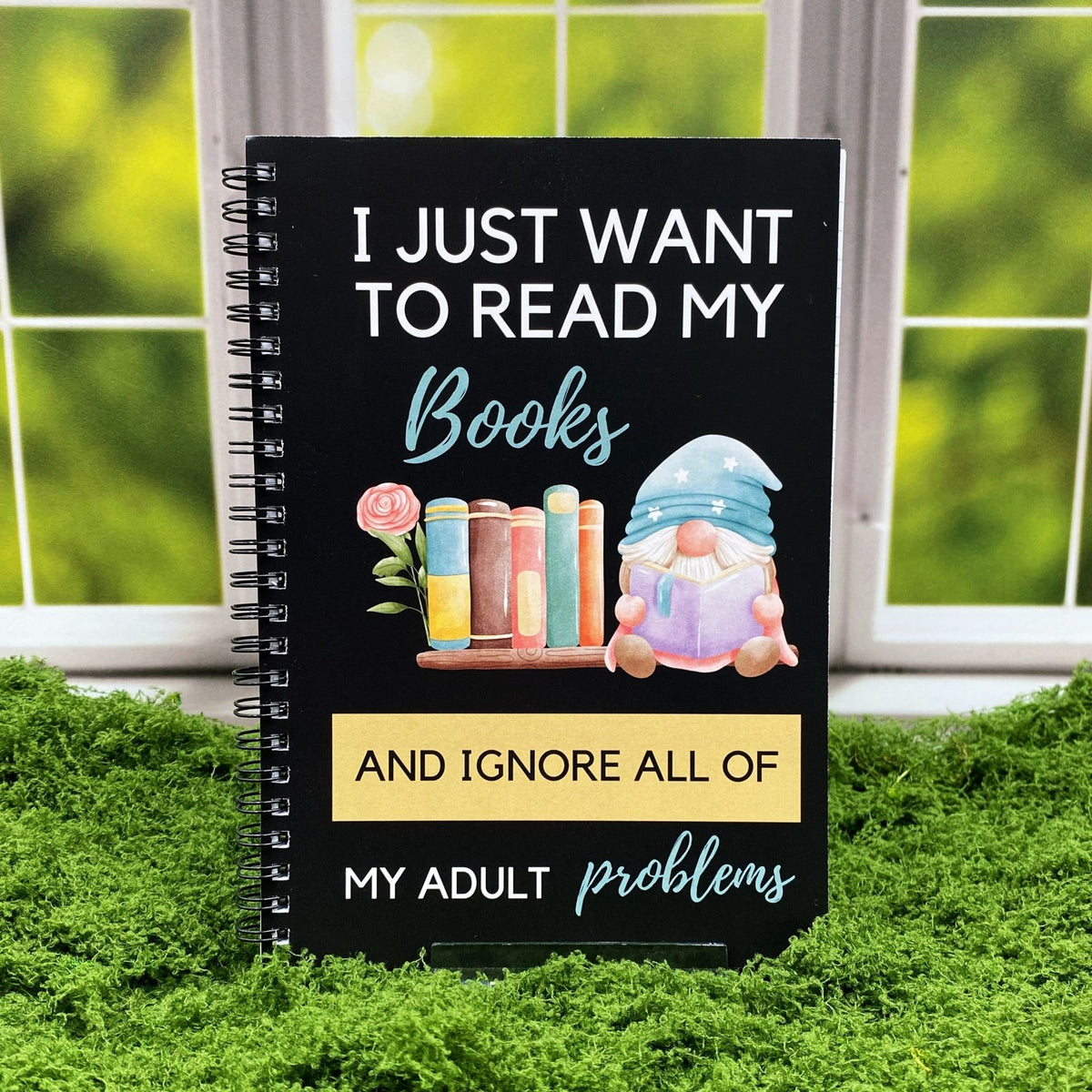 Spiral-bound black notebook reading 'I just want to read my Books and ignore all of my adult problems' with an illustration of a gnome reading beside a stack of books and a rose, displayed on a moss mat with a window backdrop.