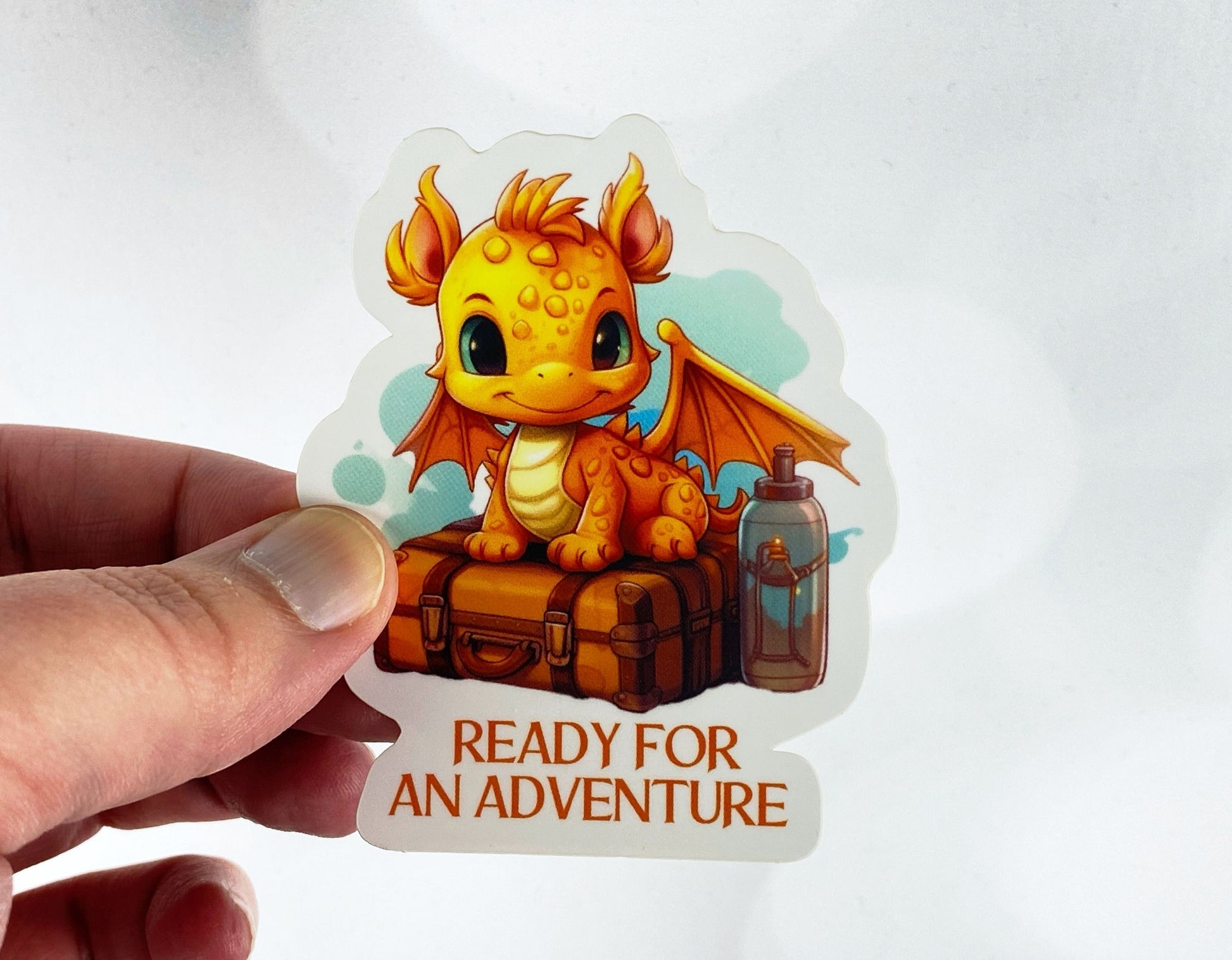 Die-cut vinyl sticker held between fingers, showing an orange baby dragon sitting on a vintage suitcase next to a lantern, above the text "Ready for an adventure," on a white background