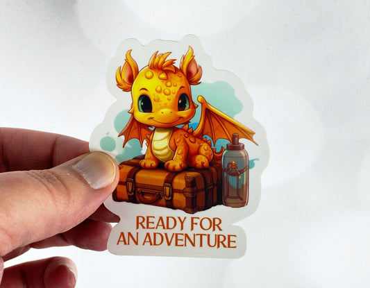 Die-cut vinyl sticker held between fingers, showing an orange baby dragon sitting on a vintage suitcase next to a lantern, above the text "Ready for an adventure," on a white background