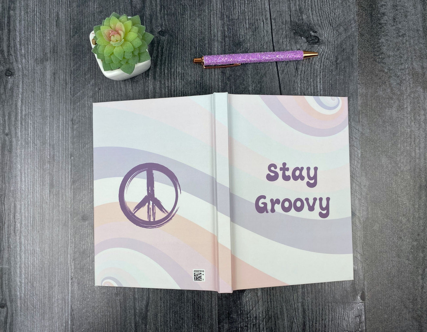 Hardcover journal open to show both covers, one with a purple peace sign on a pastel rainbow swirl background, the other reading "Stay Groovy," resting on a grey wood surface next to a glitter pen and small succulent.