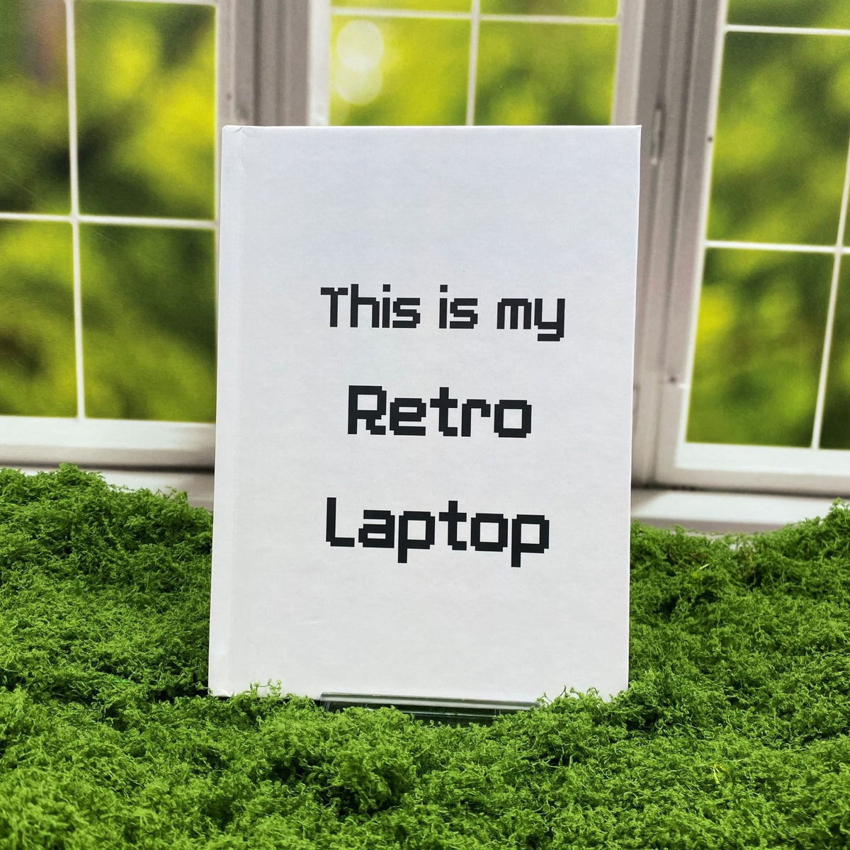 Light grey hardcover journal with pixel-style text reading 'This is my Retro Laptop,' displayed on a moss mat with a window backdrop.