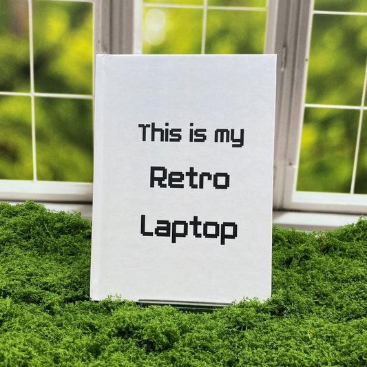 Light grey hardcover journal with pixel-style text reading 'This is my Retro Laptop,' displayed on a moss mat with a window backdrop.