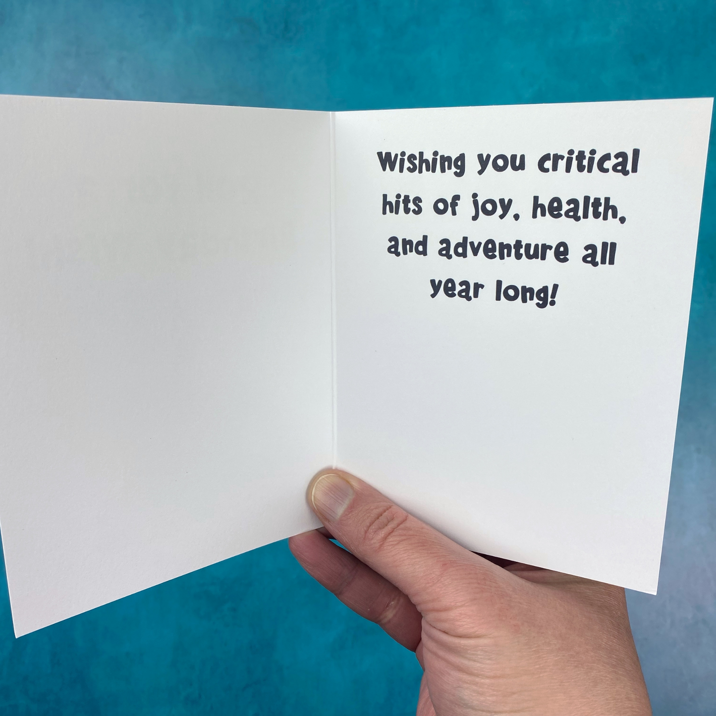 A hand holds open the greeting card, showing the printed message "Wishing you critical hits of joy, health, and adventure all year long!" against a teal background.