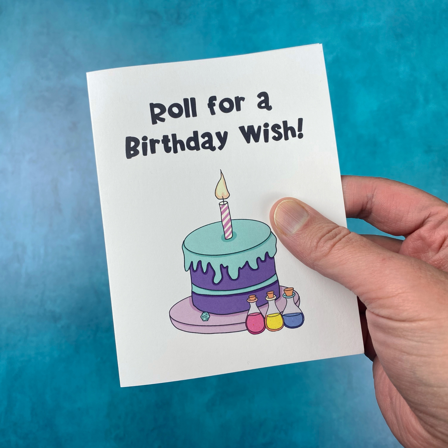 A hand holds a greeting card that reads "Roll for a Birthday Wish!" with an illustration of a birthday cake, a lit candle, a d20, and three potion bottles, against a teal background.
