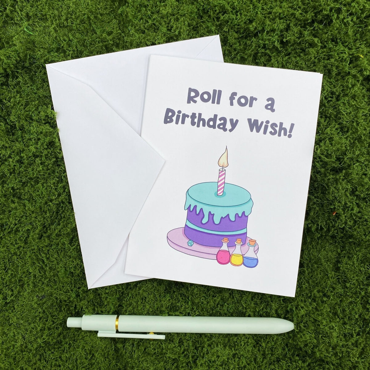 "Roll for a Birthday Wish" greeting card lying on a white envelope on grass, with a sage green pen beside it for scale.