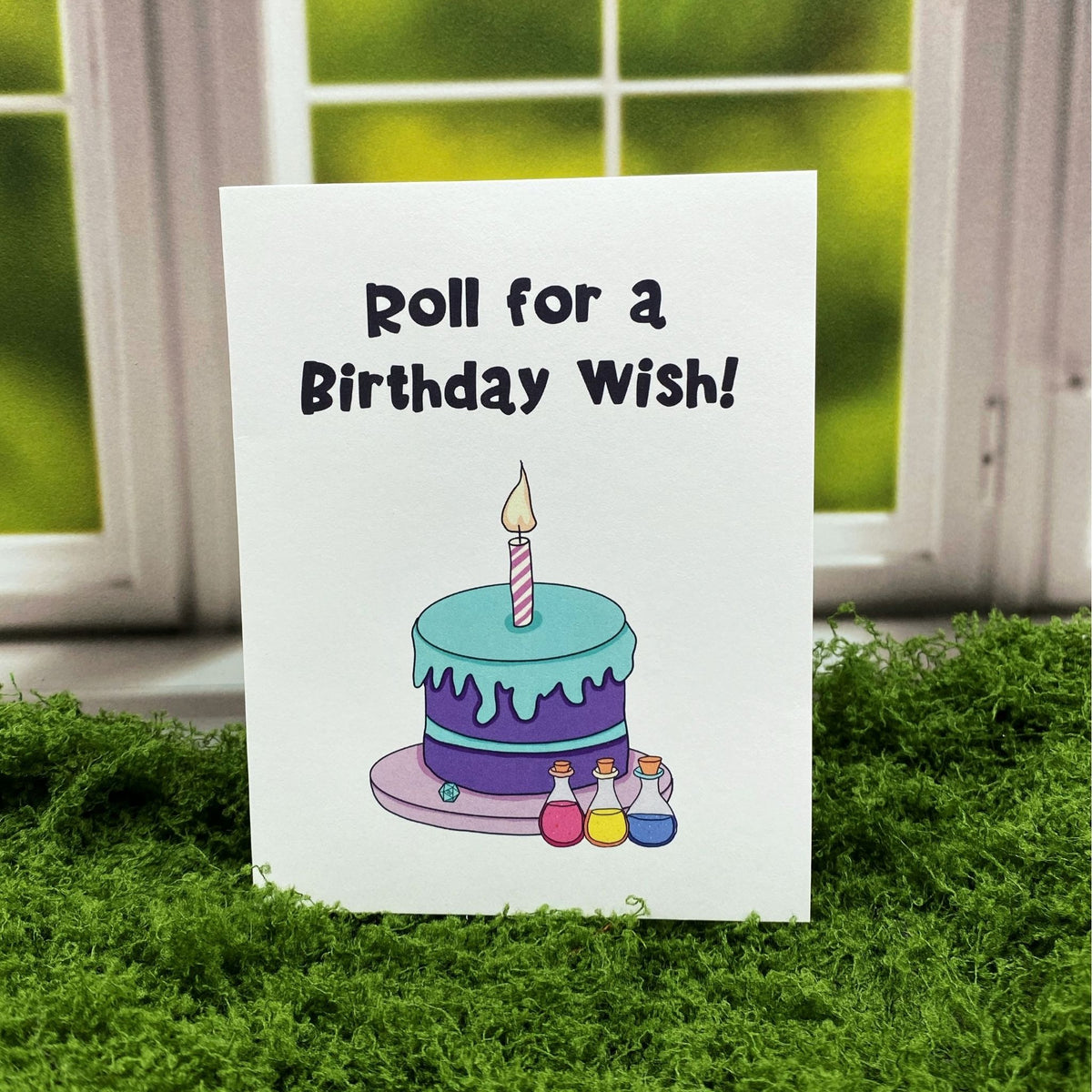 "Roll for a Birthday Wish" tabletop RPG-themed greeting card standing upright outdoors, featuring an illustrated fantasy birthday cake with a lit candle and three colorful potion bottles.