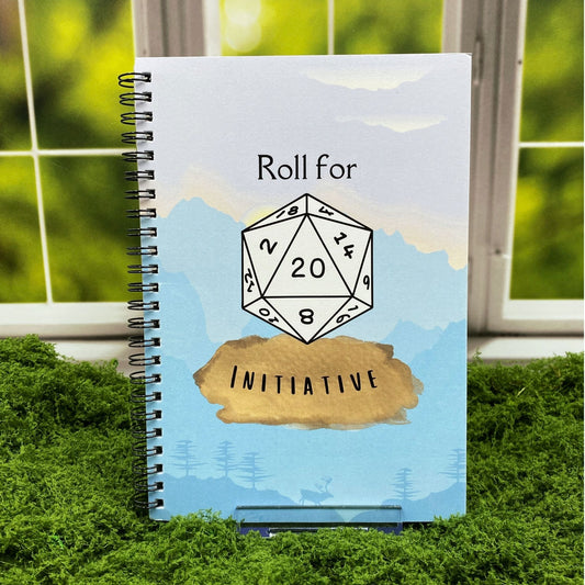 Spiral-bound 'Roll for Initiative' notebook with a d20 illustration showing a natural 20, set against a watercolor mountain and forest cover, displayed on moss with a window backdrop.
