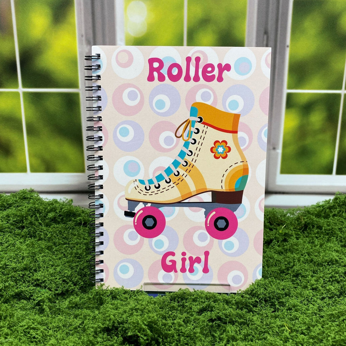 Spiral-bound notebook with a retro circle pattern and an illustrated yellow roller skate with pink wheels, reading 'Roller Girl,' displayed on a moss mat with a window backdrop.