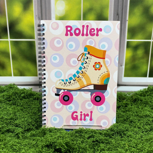 Spiral-bound notebook with a retro circle pattern and an illustrated yellow roller skate with pink wheels, reading 'Roller Girl,' displayed on a moss mat with a window backdrop.