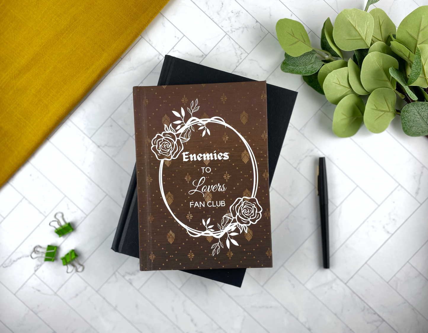 Hardcover journal in brown with a gold damask pattern, a white floral wreath, and the words "Enemies to Lovers Fan Club," resting on a black book with green binder clips, and a black pen nearby.