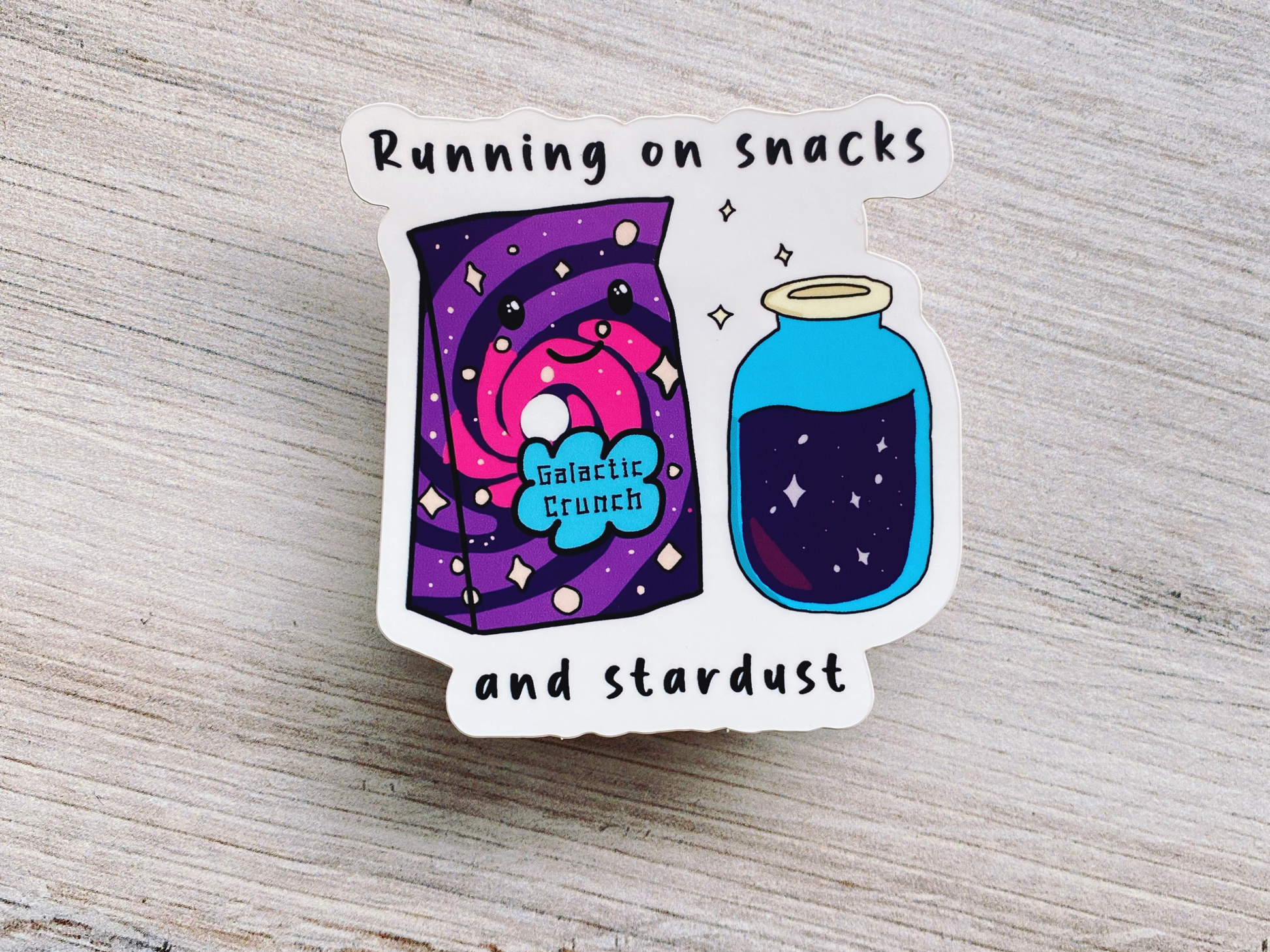 Die-cut vinyl sticker showing a bag of "Galactic Crunch" cereal next to a jar of blue stardust, with the text "Running on snacks and stardust," on a light wood background