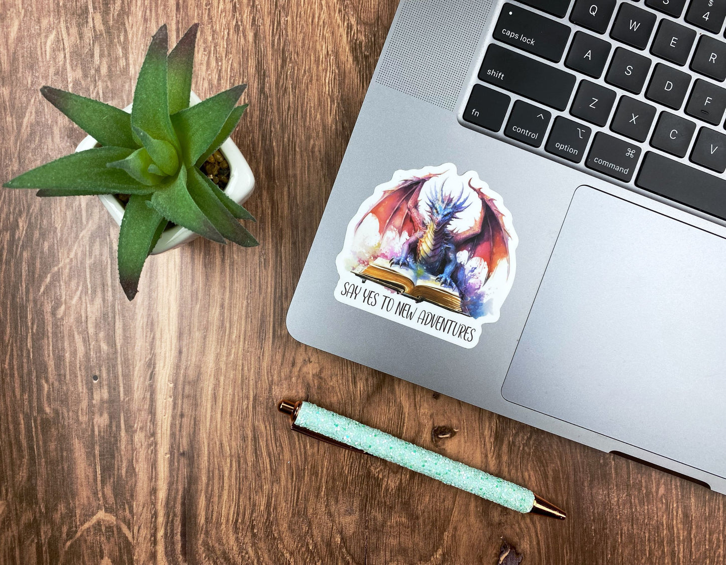 Watercolor dragon sticker applied to the corner of a silver laptop next to a succulent and a glitter pen on a wood desk