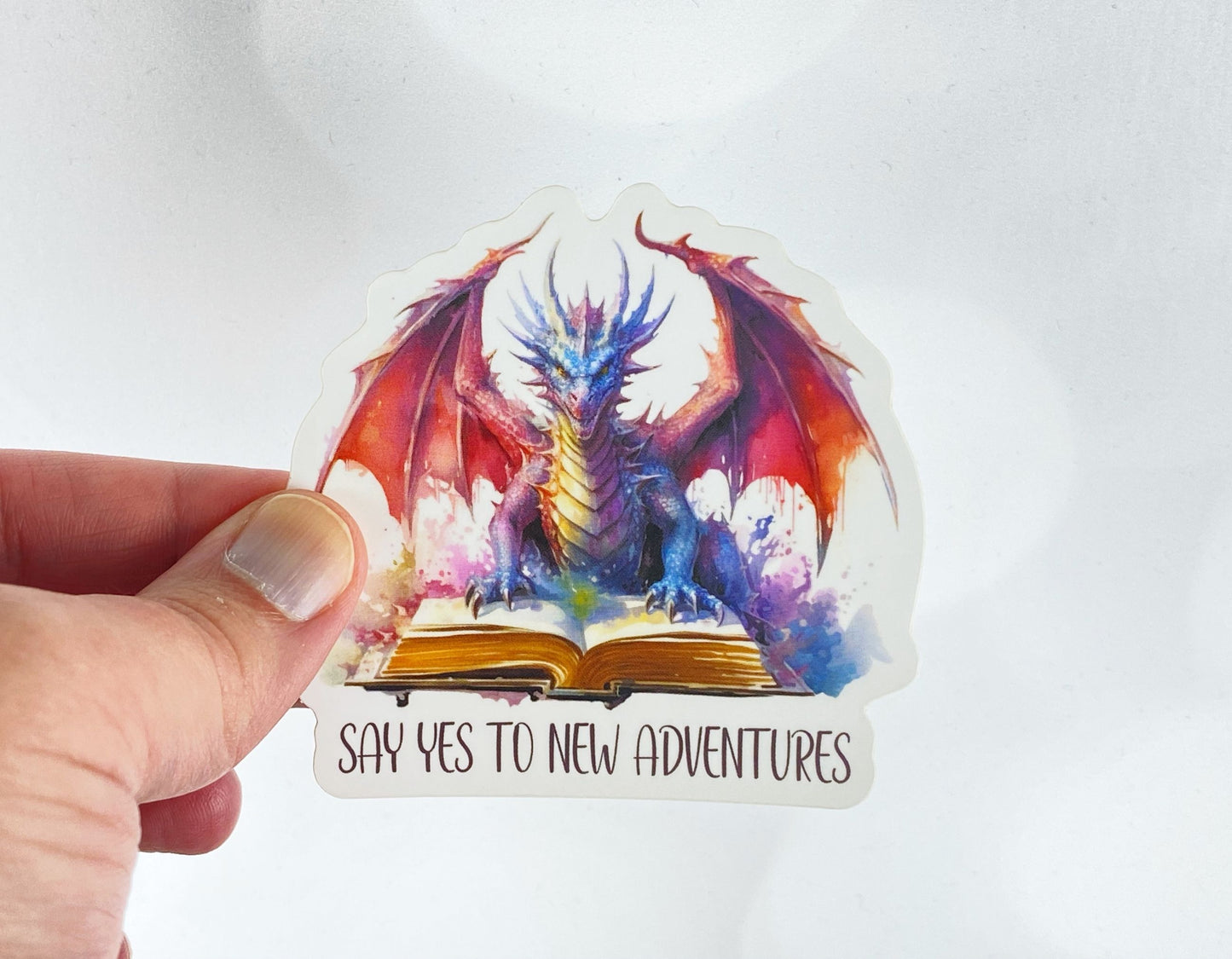 Die-cut vinyl sticker held between fingers, showing a red and blue watercolor dragon perched on an open book above the text "Say yes to new adventures," on a white background
